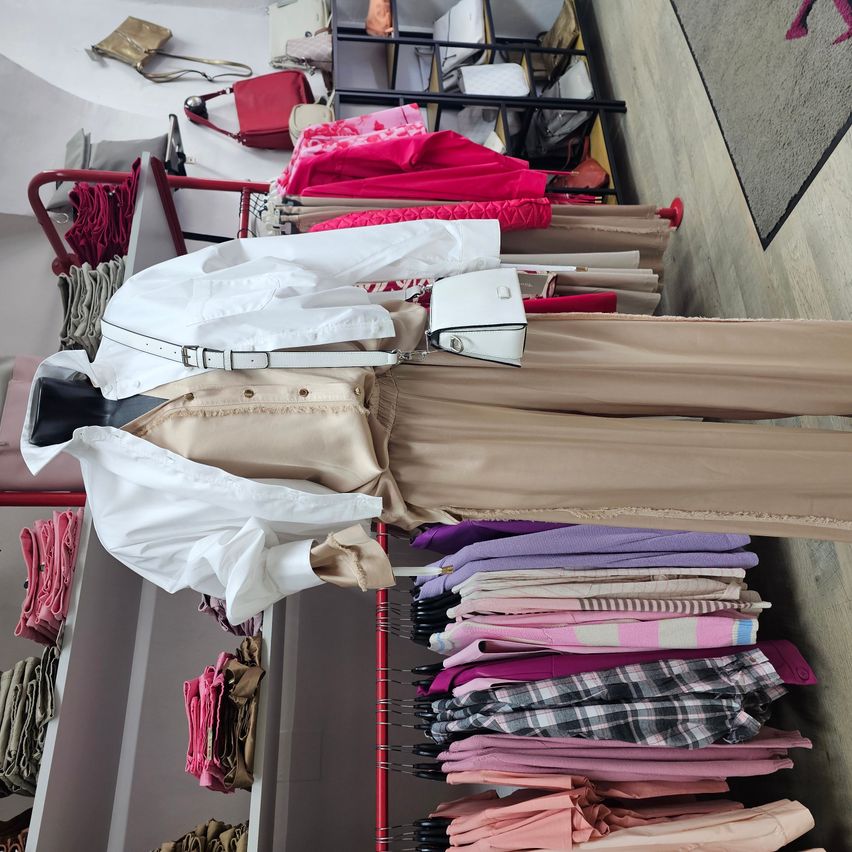 A clothing store displays a white shirt, beige pants, and a white purse on a mannequin with clothes and handbags in the background.