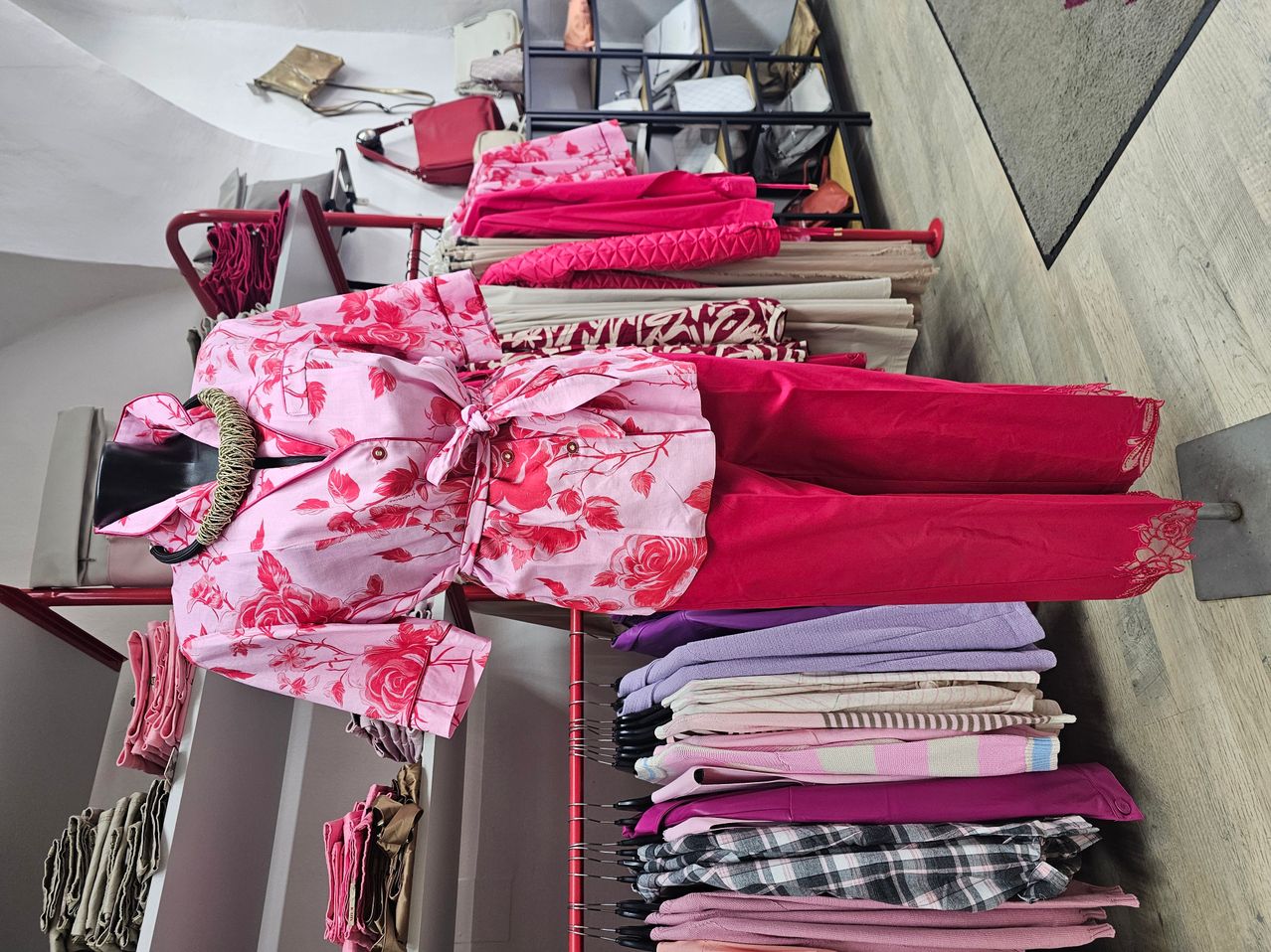 A clothing store displays a variety of clothes, including a pink floral blouse, red pants, and other garments on hangers and shelves.