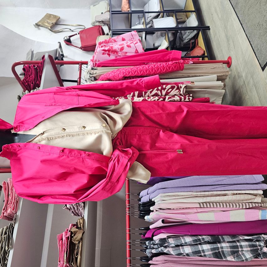 A clothing store with racks of various pink clothing, a beige top, and a pink jacket. The racks are on a red stand, and the background shows more pink and white clothing.