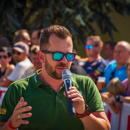 Ein Mann in einem grünen Hemd und Sonnenbrille spricht in ein Mikrofon, während eine Menge ihn im Hintergrund beobachtet.