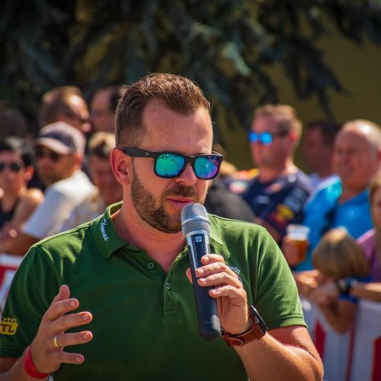 Ein Mann in einem grünen Hemd und Sonnenbrille spricht in ein Mikrofon, während eine Menge ihn im Hintergrund beobachtet.