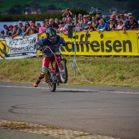 Ein Motorradfahrer in Helm und Rennausrüstung führt ein Radstandmanöver auf einer Rennstrecke durch, während eine Zuschauermenge hinter einem gelben Banner zuschaut.