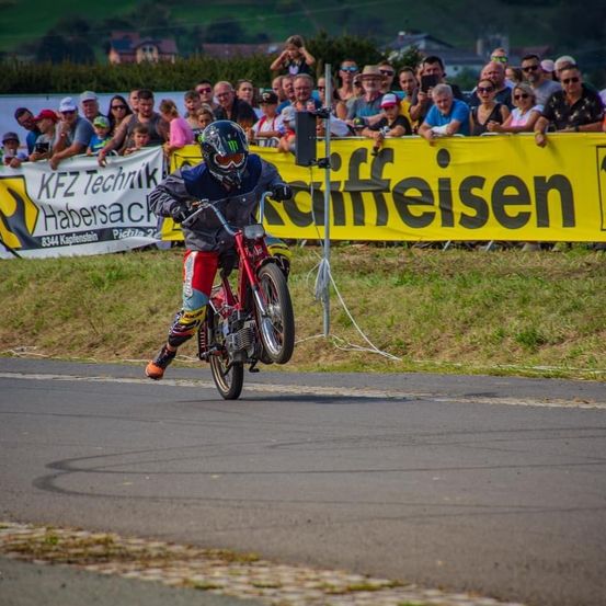 Ein Motorradfahrer in Helm und Rennausrüstung führt ein Radstandmanöver auf einer Rennstrecke durch, während eine Zuschauermenge hinter einem gelben Banner zuschaut.