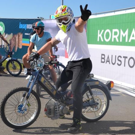Ein Mann mit Helm und Handschuhen fährt einen blauen Roller. Er zeigt den Daumen nach oben. Ein anderer Mann auf einem Roller fährt hinter ihm. Sie stehen vor einem Banner, auf dem Korma Bauston steht.