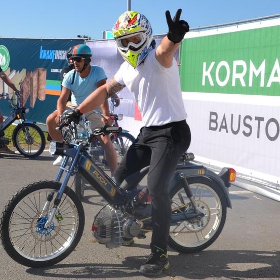 Ein Mann mit Helm und Handschuhen fährt einen blauen Roller. Er zeigt den Daumen nach oben. Ein anderer Mann auf einem Roller fährt hinter ihm. Sie stehen vor einem Banner, auf dem Korma Bauston steht.