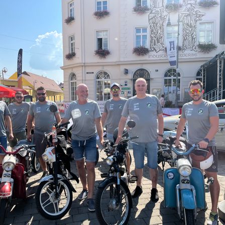 Sechs Männer stehen neben Vintage-Rollern und Motorrädern auf einer Kopfsteinpflasterstraße vor einem weißen Gebäude mit dekorativen Elementen.