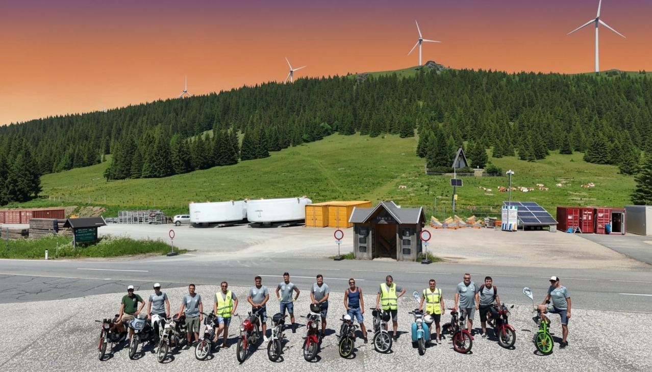 A group of people in safety vests stand in front of a mountain with wind turbines, posing for a photo. Behind them, there are storage boxes, a small building, and a road with traffic signs.