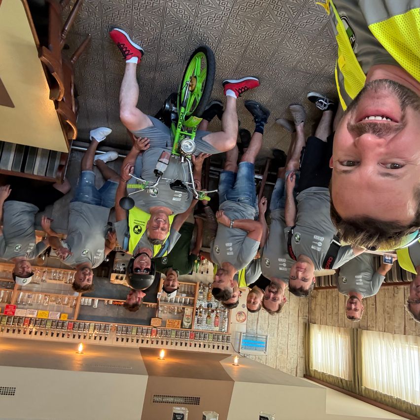 A group of men in gray shirts and reflective vests are upside down, holding onto a green bicycle with a black seat and red wheels. They are in a room with wooden walls and shelves filled with items.