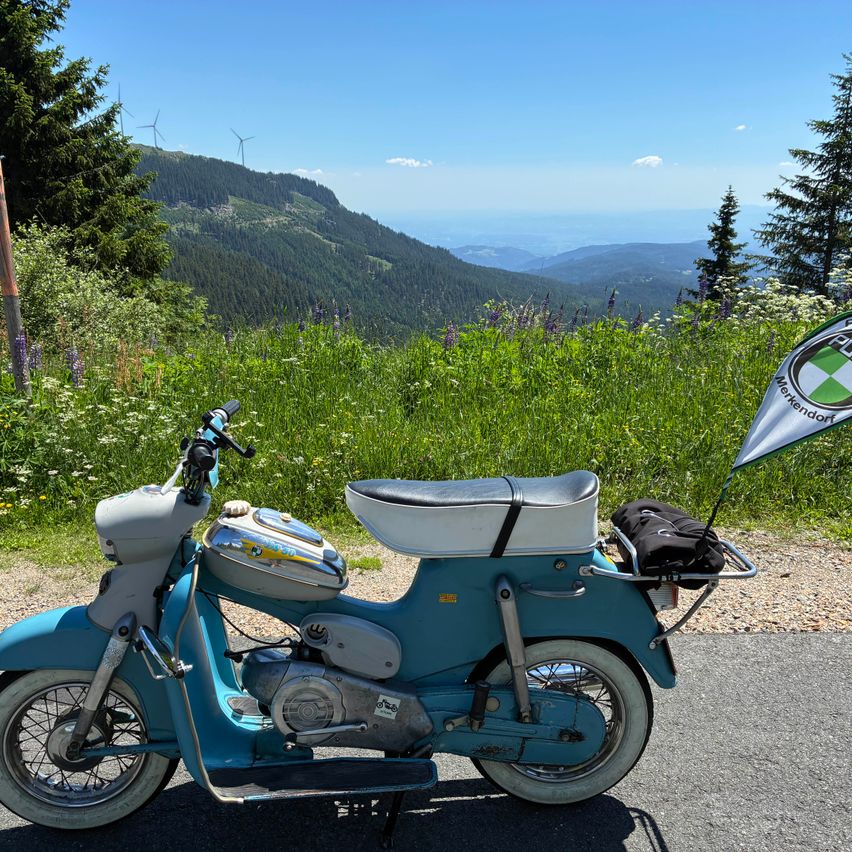A vintage blue moped is parked on the side of a mountain road, with a scenic view of mountains and a flag in the background.