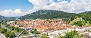 Eine Luftaufnahme einer Stadt mit vielen Gebäuden, Bäumen und Bergen. Ein Fluss und eine Brücke sind im Vordergrund.
