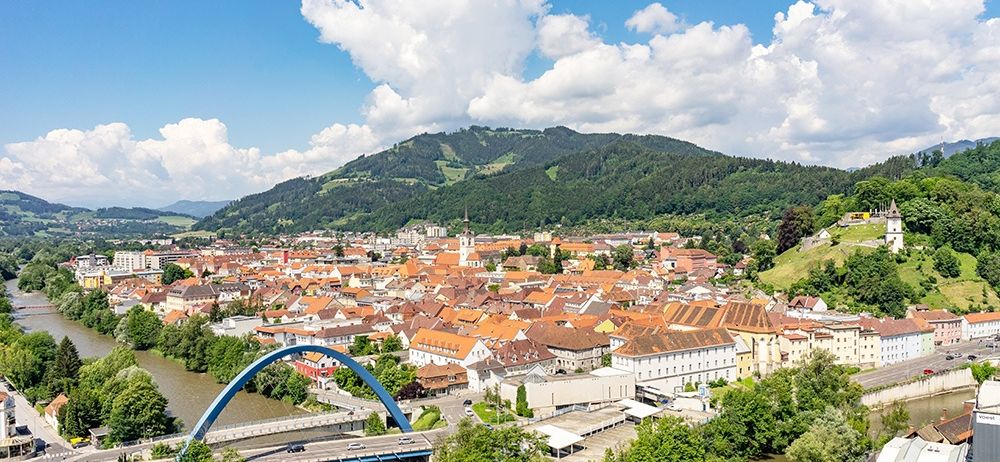 Eine Luftaufnahme einer Stadt mit vielen Gebäuden, Bäumen und Bergen. Ein Fluss und eine Brücke sind im Vordergrund.