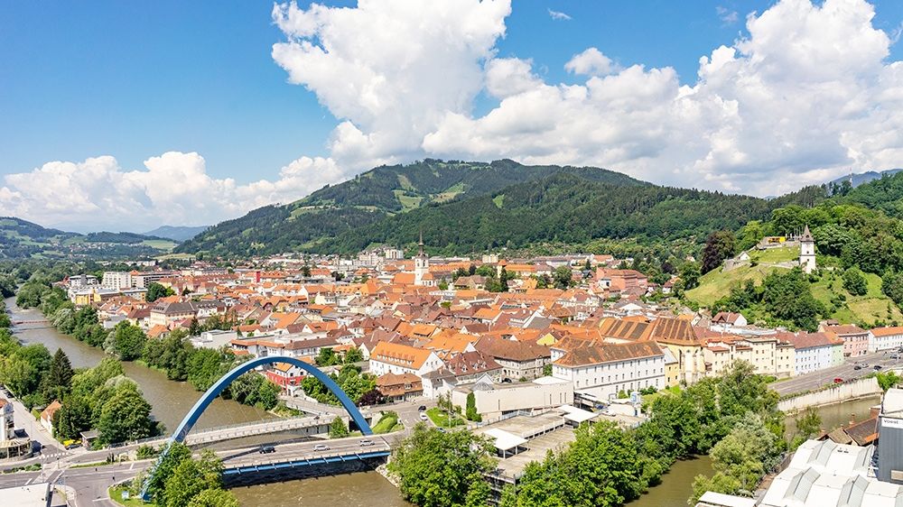 Eine Luftaufnahme einer Stadt mit vielen Gebäuden, Bäumen und Bergen. Ein Fluss und eine Brücke sind im Vordergrund.