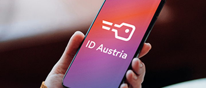 Eine Person hält ein Smartphone mit der ID Austria-App, die auf einem lila und rosa Hintergrund mit einem weißen Symbol angezeigt wird.