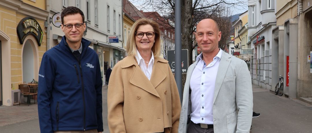 Drei Personen, zwei Männer und eine Frau, stehen auf einer Straße, die von Gebäuden gesäumt ist. Die Frau trägt eine Brille und einen beigen Mantel, während die Männer eine blaue Jacke und einen hellgrauen Anzug tragen. Hinter ihnen steht ein blattloser Baum, und auf beiden Seiten der Straße sind Gebäude zu sehen.