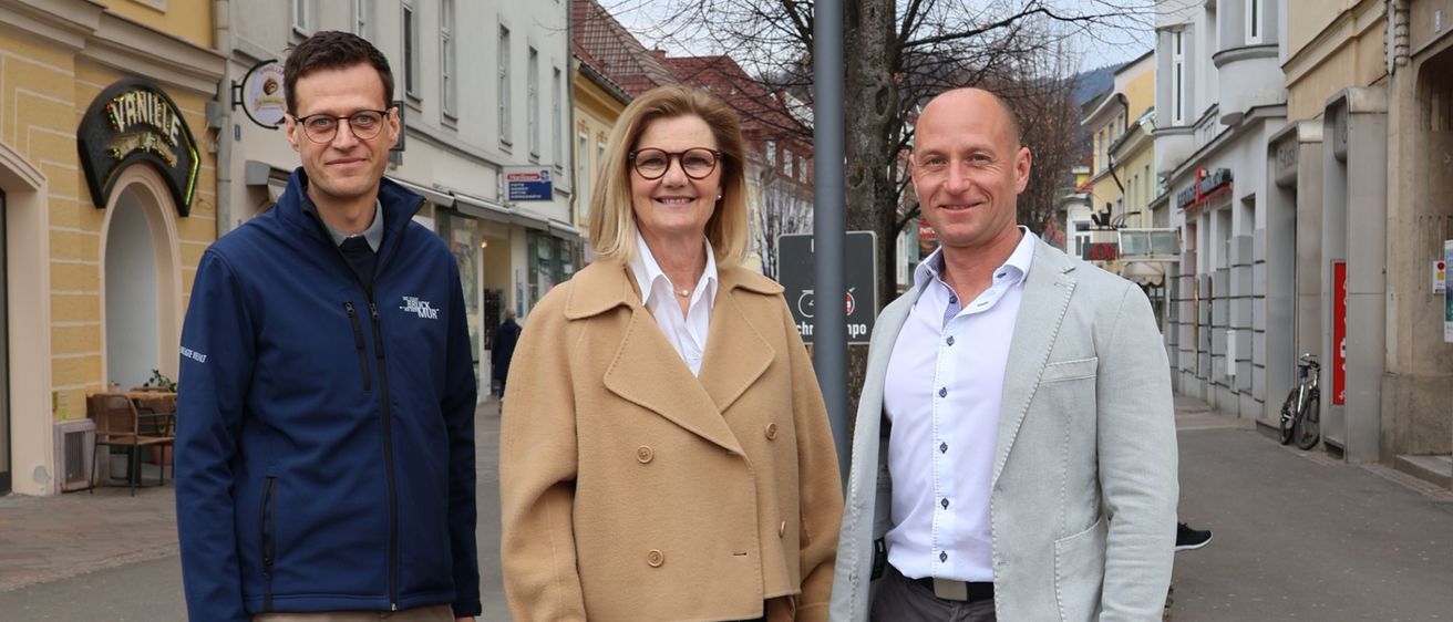 Drei Personen, zwei Männer und eine Frau, stehen auf einer Straße, die von Gebäuden gesäumt ist. Die Frau trägt eine Brille und einen beigen Mantel, während die Männer eine blaue Jacke und einen hellgrauen Anzug tragen. Hinter ihnen steht ein blattloser Baum, und auf beiden Seiten der Straße sind Gebäude zu sehen.