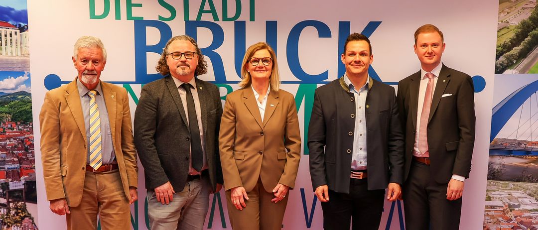 Fünf Personen, die feierlich gekleidet sind, stehen auf einem roten Teppich vor einem Hintergrund mit dem Text 'Die Stadt Bruck'.