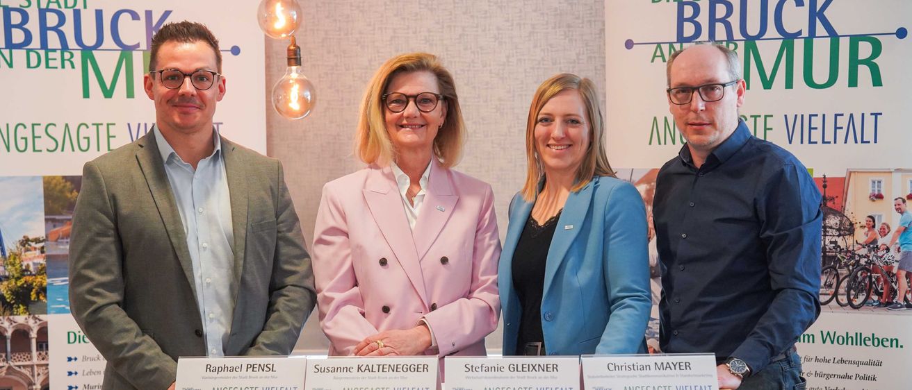 Vier Personen stehen lächelnd hinter einem Podium und posieren für die Kamera. Sie tragen alle formelle Kleidung. Zwei Frauen tragen Blazer, während die Männer Anzüge tragen. Jede Person hat einen Namensschild mit ihrem Namen und dem Text 'angesagt Vielfalt'.