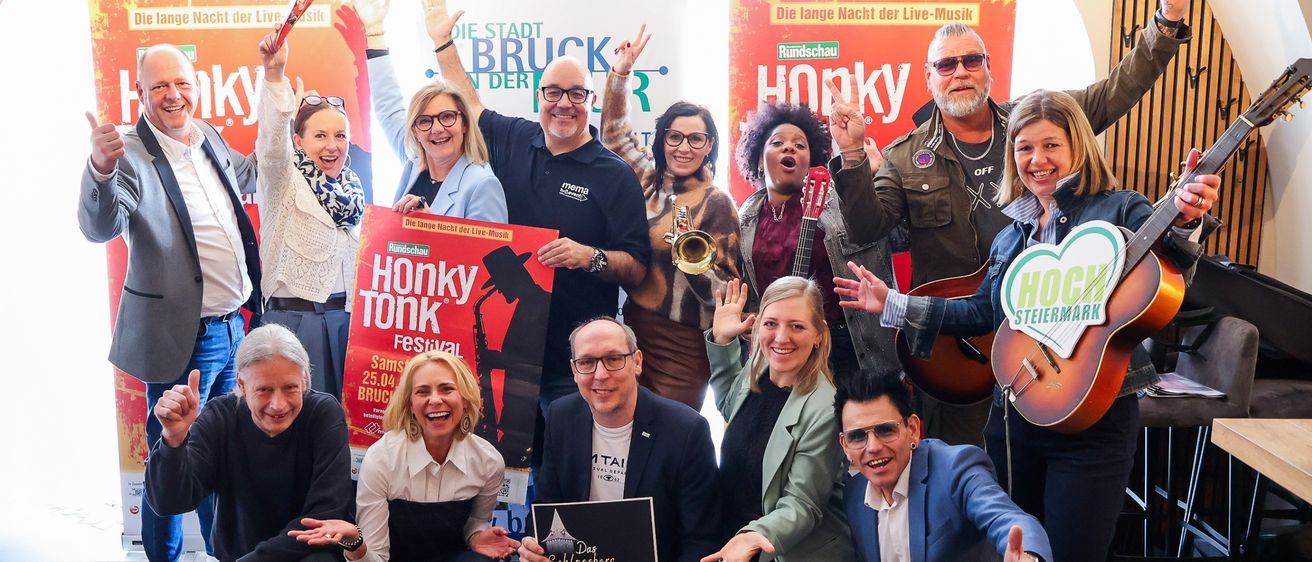Eine Gruppe von Menschen posiert für ein Foto mit Musikinstrumenten und einem Plakat, das Honky Tonk Festival liest. Einige halten Gitarren, Trompeten und ein Schild, und sie alle lächeln und schauen in die Kamera.