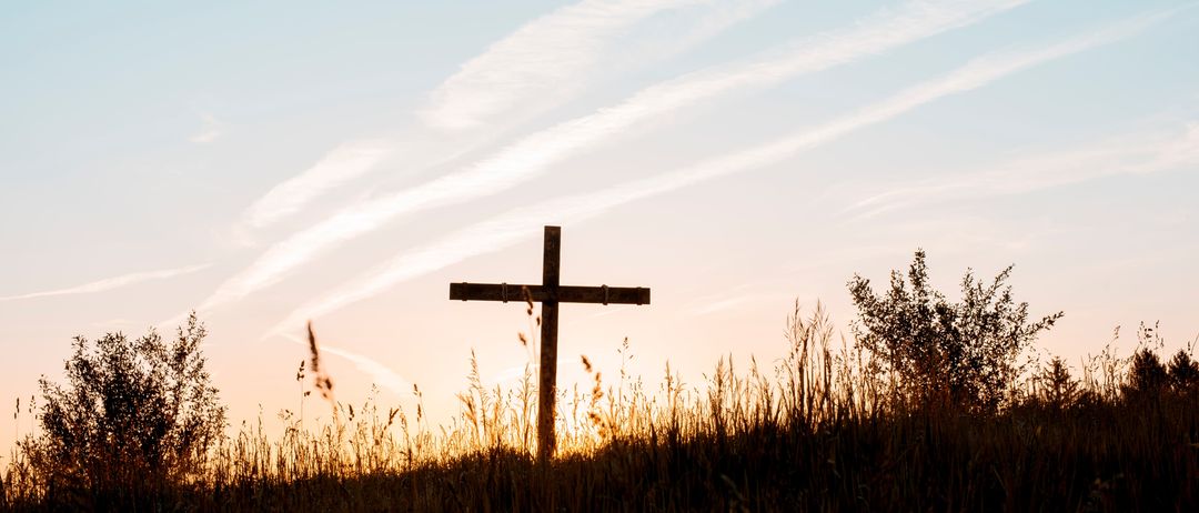 Ein hölzernes Kreuz steht auf einer grasbewachsenen Wiese bei Sonnenuntergang und wirft einen langen Schatten auf den Himmel.