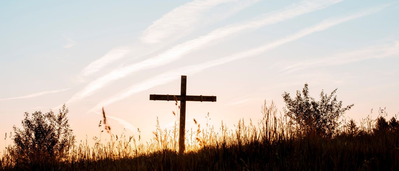 Ein hölzernes Kreuz steht auf einer grasbewachsenen Wiese bei Sonnenuntergang und wirft einen langen Schatten auf den Himmel.