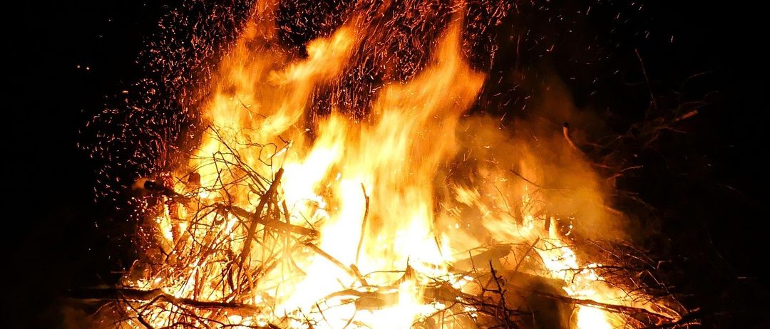 Ein Lagerfeuer brennt intensiv in der Nacht und wirft flackernde Schatten. Funken fliegen in die Luft. Die Flammen tanzen wild umher, umgeben von einem Haufen Holz.