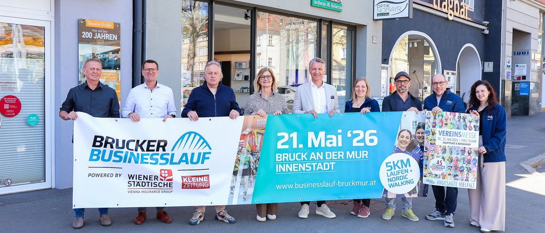 Eine Gruppe von Menschen steht vor einem Gebäude und hält Plakate in der Hand. Sie scheinen eine Veranstaltung zu bewerben. Das Banner trägt den Text Brucker Businesslauf, Bruck an der Mur Innenstadt, mit einer Website.