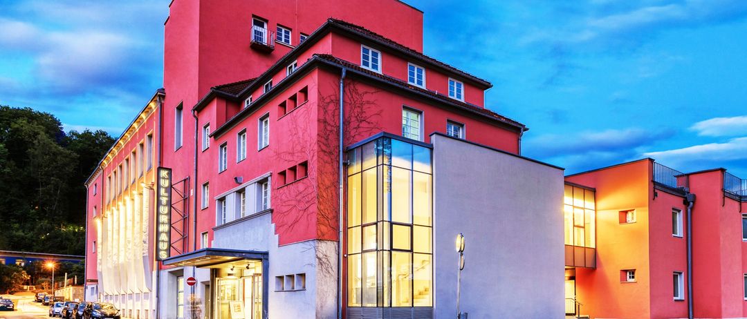 Ein modernes rotes Gebäude mit Glasfenstern ist nachts beleuchtet. Ein Auto ist auf der Straßenseite geparkt.