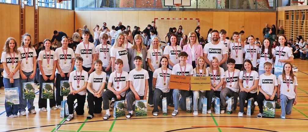 Eine Gruppe junger Athleten in weißen Hemden und Medaillen posiert für ein Foto in einer Turnhalle. Einige halten Pokale und Taschen. Im Hintergrund befindet sich eine große Kletterwand und ein Basketballkorb.