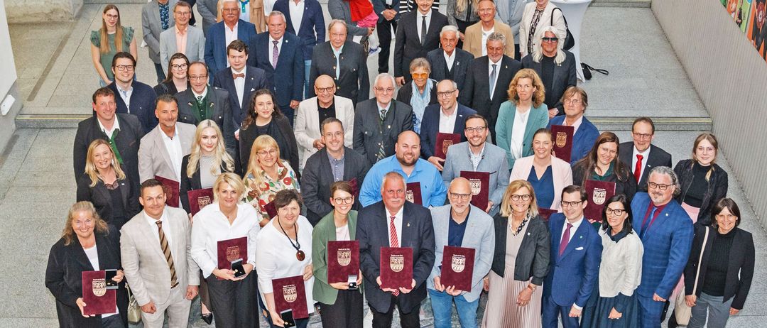 Ein Gruppenfoto von Personen in formeller Kleidung, möglicherweise bei einer Preisverleihung, einige halten Auszeichnungen. Eine Frau hält ein Kind, andere stehen hinter Tischen.