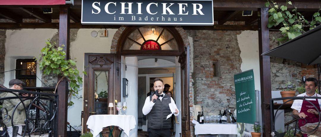 Ein Mann mit Mikrofon steht vor einem Tisch am Eingang des Schicker im Baderhaus, einem Restaurant. Flaschen und Weingläser stehen auf dem Tisch. Töpfe mit Pflanzen befinden sich auf beiden Seiten des Eingangs.