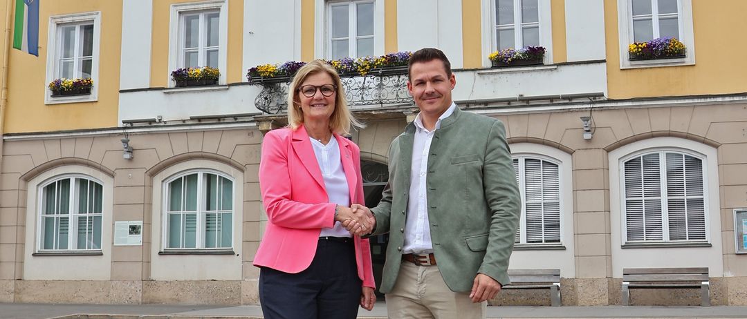 Ein Mann und eine Frau stehen lächelnd vor dem Rathaus-Gebäude mit einer Uhr und dekorativen Fensterbänken.