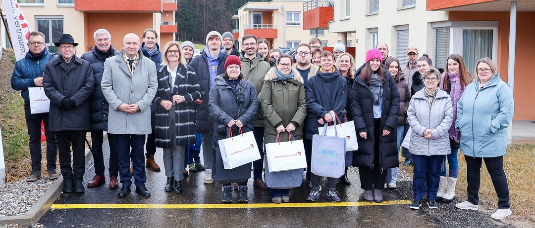 Eine Gruppe von Menschen steht auf einer nassen Straße, Einkaufstüten in der Hand. Sie stehen vor einigen Gebäuden mit Balkonen.