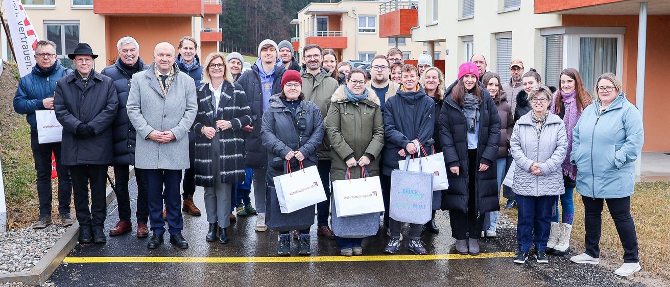 Eine Gruppe von Menschen steht auf einer nassen Straße, Einkaufstüten in der Hand. Sie stehen vor einigen Gebäuden mit Balkonen.