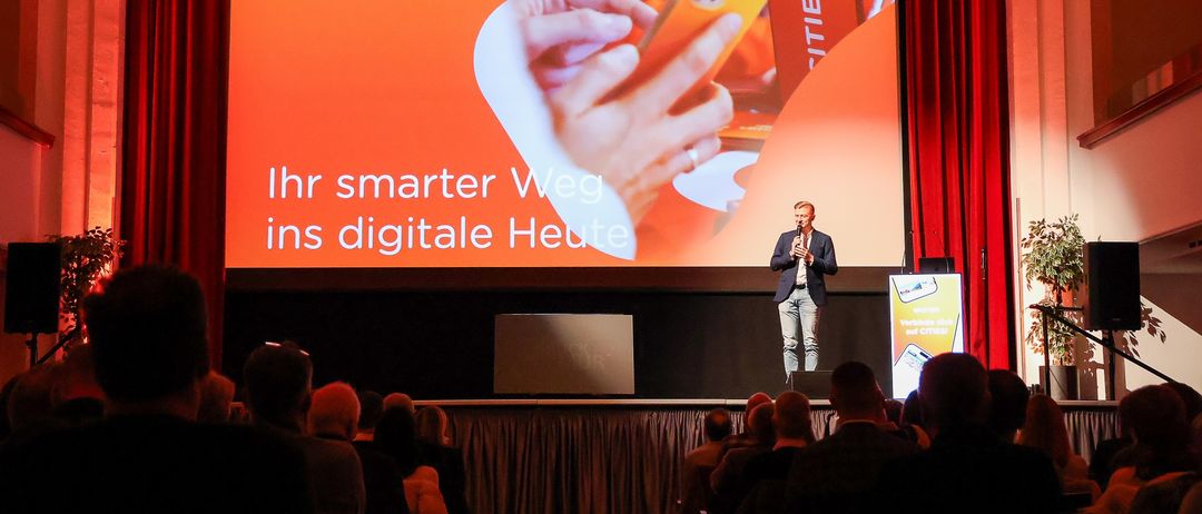 Ein Mann steht auf der Bühne mit einem Mikrofon und spricht vor einem Publikum. Hinter ihm zeigt eine große Leinwand eine Botschaft auf Deutsch, 'Ihr smarter Weg ins digitale Heute.'