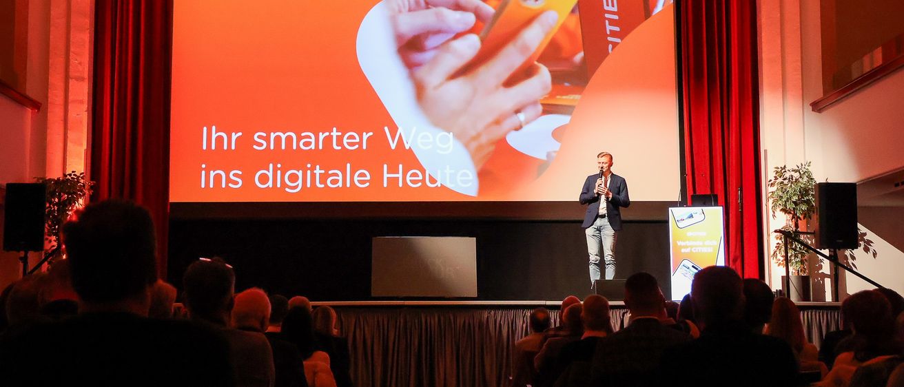 Ein Mann steht auf der Bühne mit einem Mikrofon und spricht vor einem Publikum. Hinter ihm zeigt eine große Leinwand eine Botschaft auf Deutsch, 'Ihr smarter Weg ins digitale Heute.'