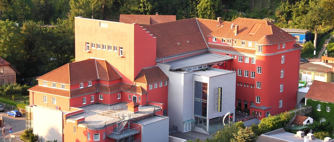 Eine Vogelperspektive auf ein großes mehrstöckiges Gebäude mit roten Wänden und braunen Dächern. Das Gebäude hat viele Fenster und ist von Bäumen umgeben.