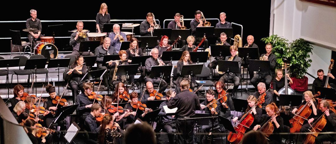 Ein vielfältiges Orchester spielt auf der Bühne mit verschiedenen Instrumenten. Der Dirigent steht davor und das Publikum sitzt davor.