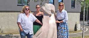 Drei Personen stehen neben einer großen Statue einer Frau, wobei eine Frau die Hand der Statue hält. Die Statue hat ein Schild mit den Aufschriften 'Freiheit' und 'Hermann Pitow'. Hinter ihnen steht ein Gebäude mit Fenstern und einem Baum.