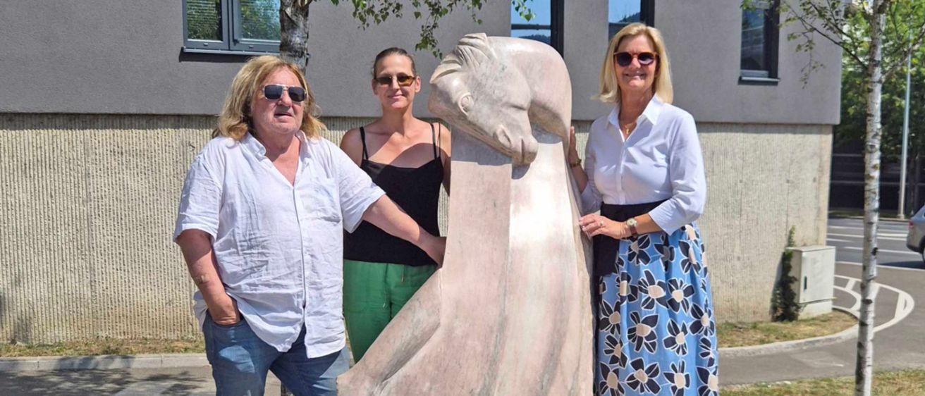 Drei Personen stehen neben einer großen Statue einer Frau, wobei eine Frau die Hand der Statue hält. Die Statue hat ein Schild mit den Aufschriften 'Freiheit' und 'Hermann Pitow'. Hinter ihnen steht ein Gebäude mit Fenstern und einem Baum.