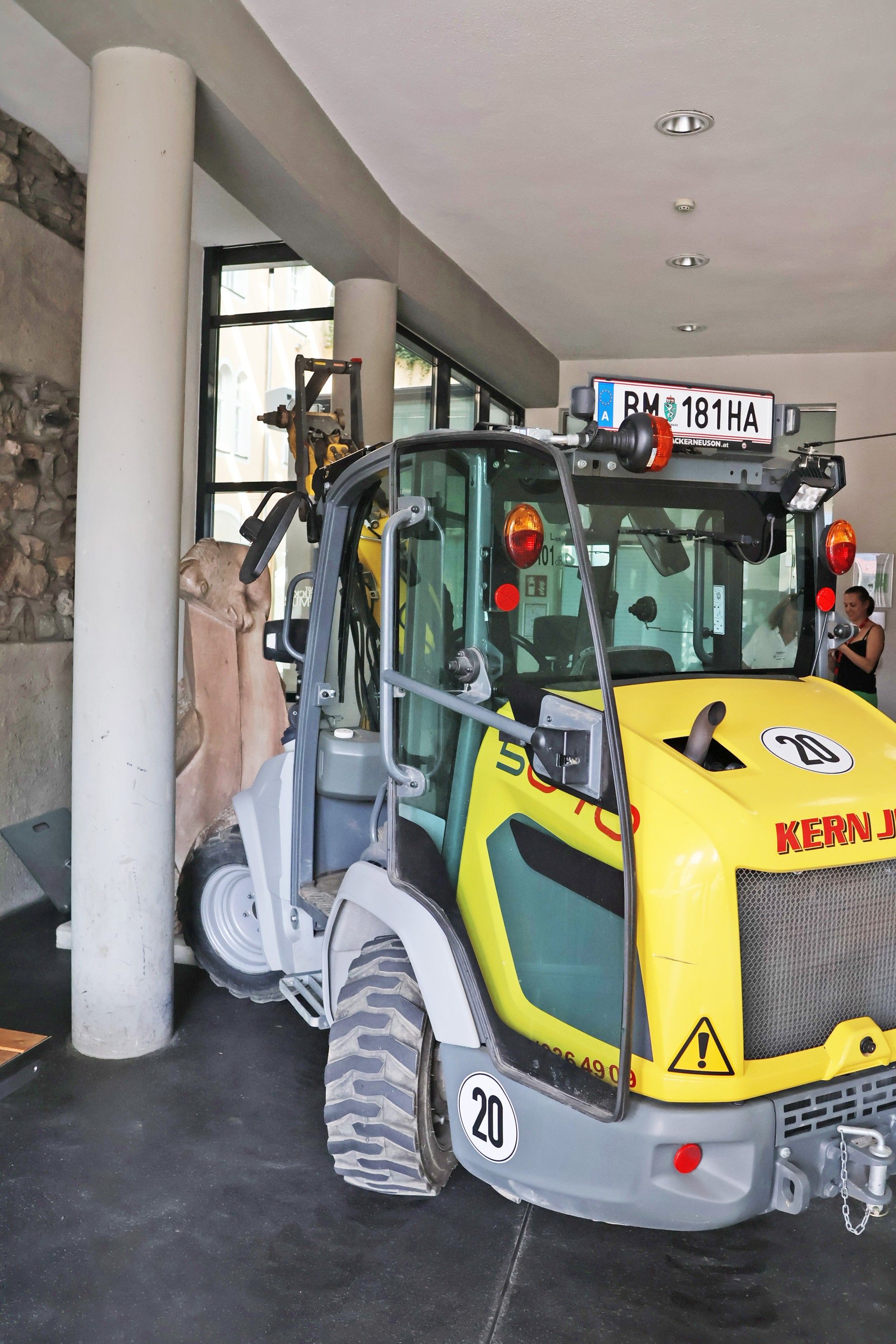 Ein gelber und grauer Baufahrzeug mit der Nummer 20 auf der Vorderseite ist in einem Gebäude geparkt. Eine Frau in einem schwarzen Tanktop steht in der Nähe.