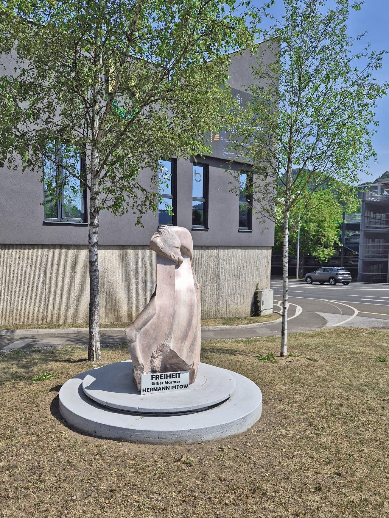 Eine Statue einer Frau steht auf einem Betonsockel in einem Park, mit einem Schild, das 'Freiheit' und 'Hermann Pitow' besagt. Die Statue ist von zwei kleinen Bäumen und einem Gebäude mit Fenstern umgeben. Ein Auto fährt auf der Straße im Hintergrund.