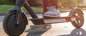 Eine Person fährt einen schwarzen Elektroroller auf einer asphaltierten Straße, trägt braune Hosen und graue Schuhe. Der Roller hat einen roten Akzent und ist an einem sonnigen Tag positioniert.