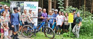 Eine Gruppe von Menschen in Fahrradausrüstung steht in einem Wald und posiert für ein Foto mit einem blauen Fahrrad im Vordergrund. Ein Schild mit dem Wort 'Start' ist rechts sichtbar.