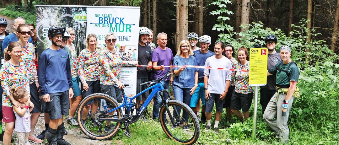 Eine Gruppe von Menschen in Fahrradausrüstung steht in einem Wald und posiert für ein Foto mit einem blauen Fahrrad im Vordergrund. Ein Schild mit dem Wort 'Start' ist rechts sichtbar.