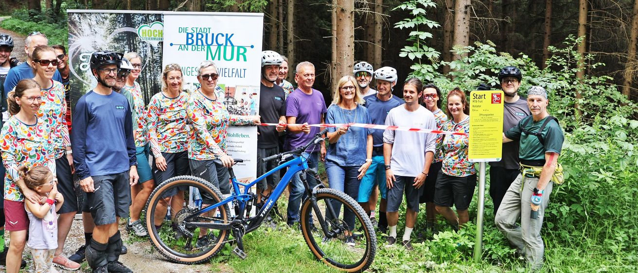 Eine Gruppe von Menschen in Fahrradausrüstung steht in einem Wald und posiert für ein Foto mit einem blauen Fahrrad im Vordergrund. Ein Schild mit dem Wort 'Start' ist rechts sichtbar.
