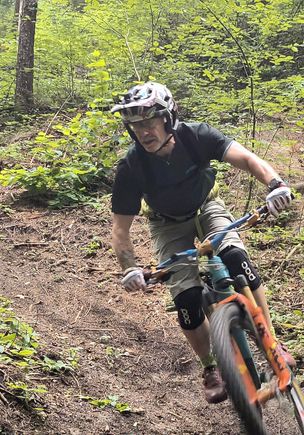 Ein Mann fährt ein Mountainbike auf einem Pfad in einem Wald. Er trägt einen Helm, eine Schutzbrille und Knieschützer.