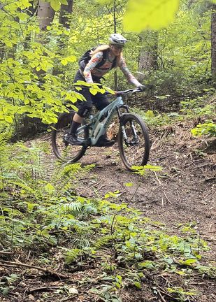 Eine Frau fährt ein Mountainbike auf einem Pfad im Wald. Sie trägt einen Helm, Handschuhe und einen Rucksack.