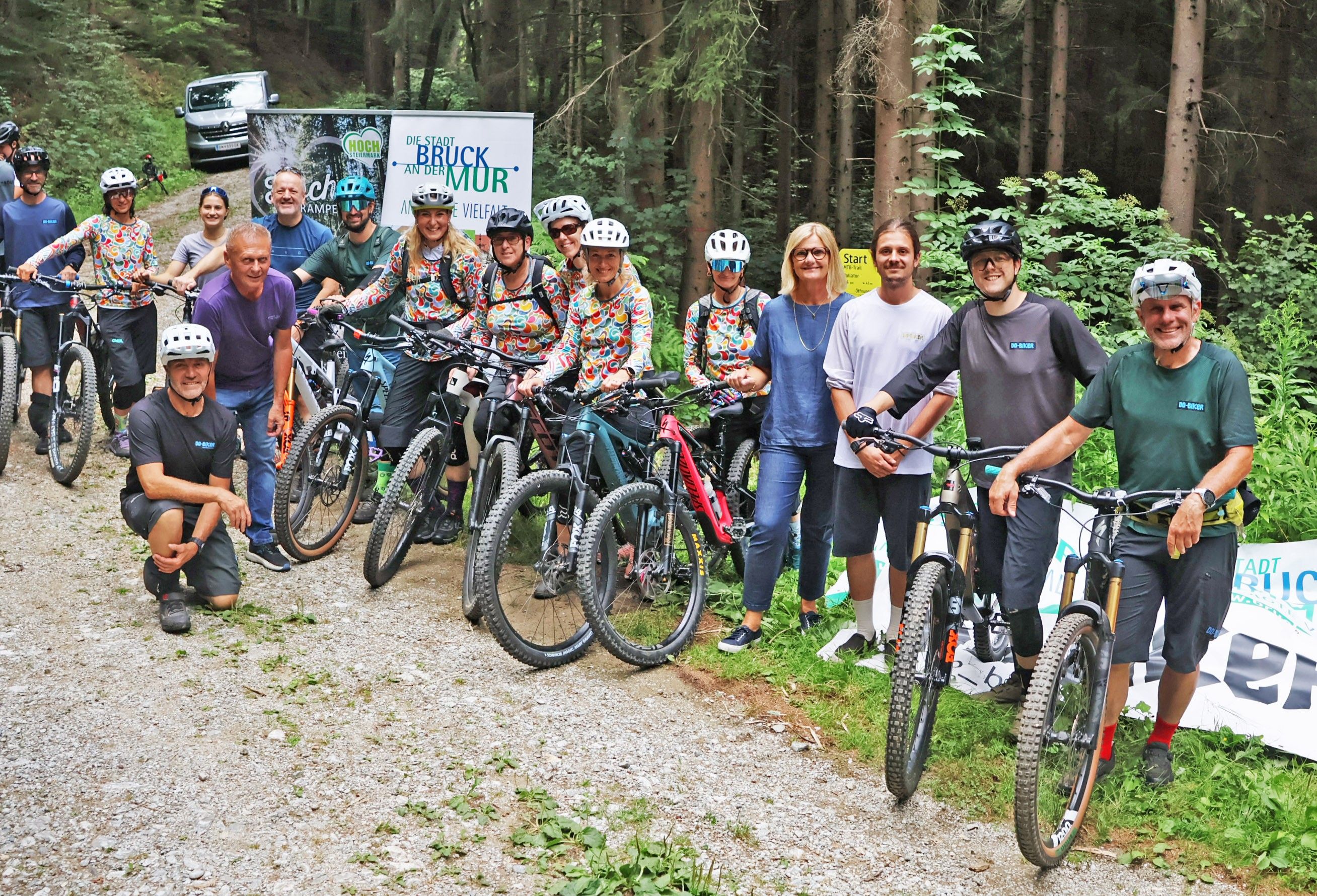 Eine Gruppe von Radfahrern in bunten Trikots und Helmen posiert für ein Foto auf einem Schotterweg im Wald. Ein Banner im Hintergrund lautet 'Die Stadt Bruck an der Mur'.