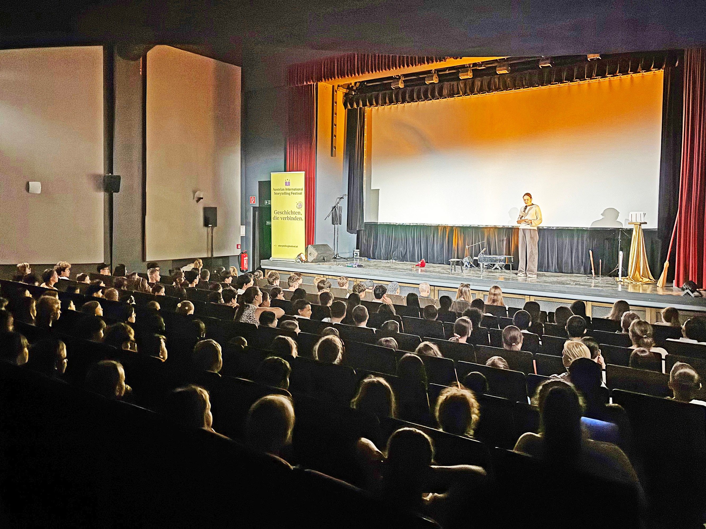 Ein Redner steht auf einer Bühne in einem Theater mit Leinwand und spricht vor einem vollen Publikum. Das Theater hat ein großes Banner links mit gelben und schwarzen Farben.