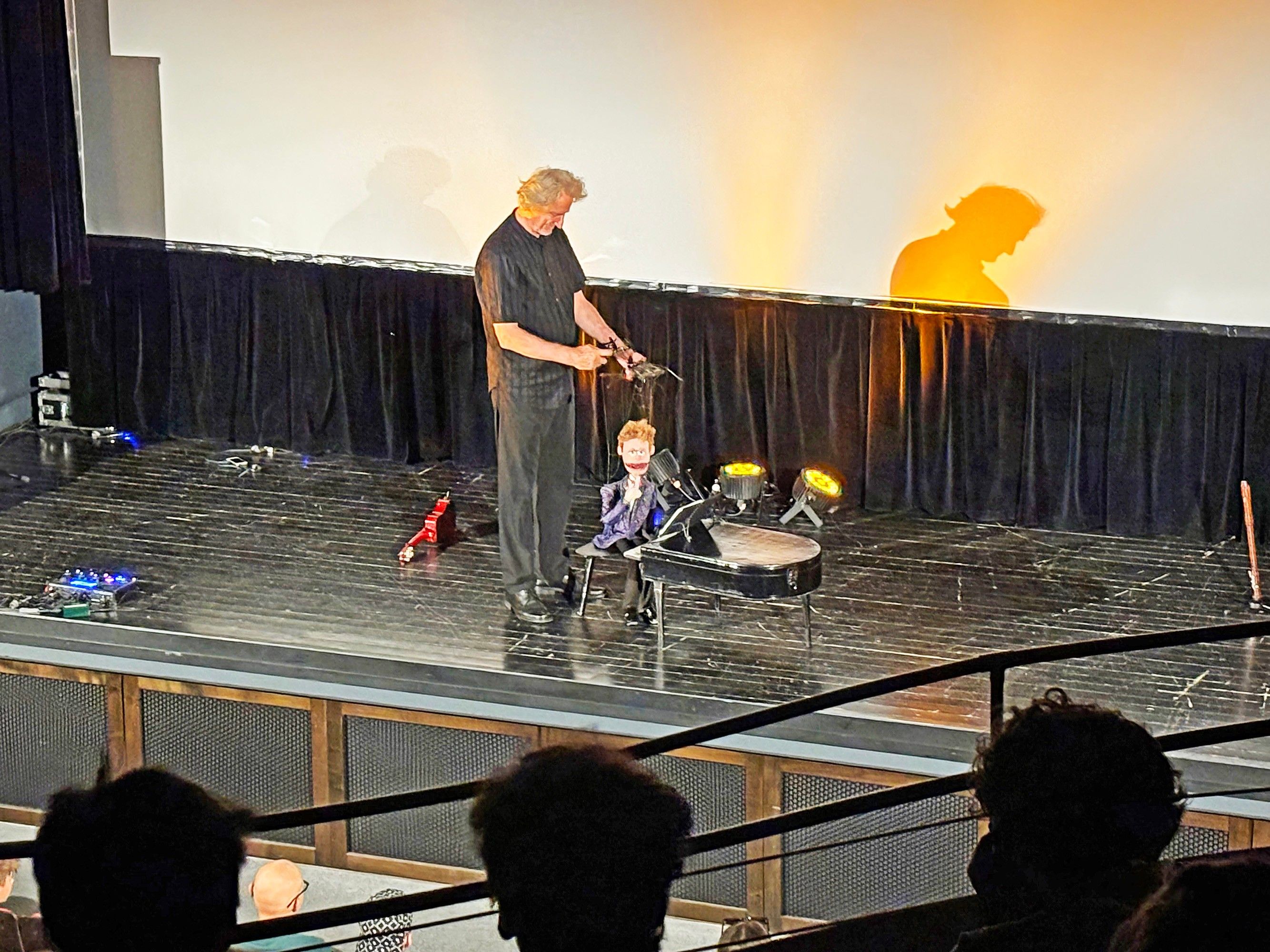 Ein Mann steht auf der Bühne mit einer Puppe vor einem Klavier unter Scheinwerfern. Das Publikum sitzt im Vordergrund.